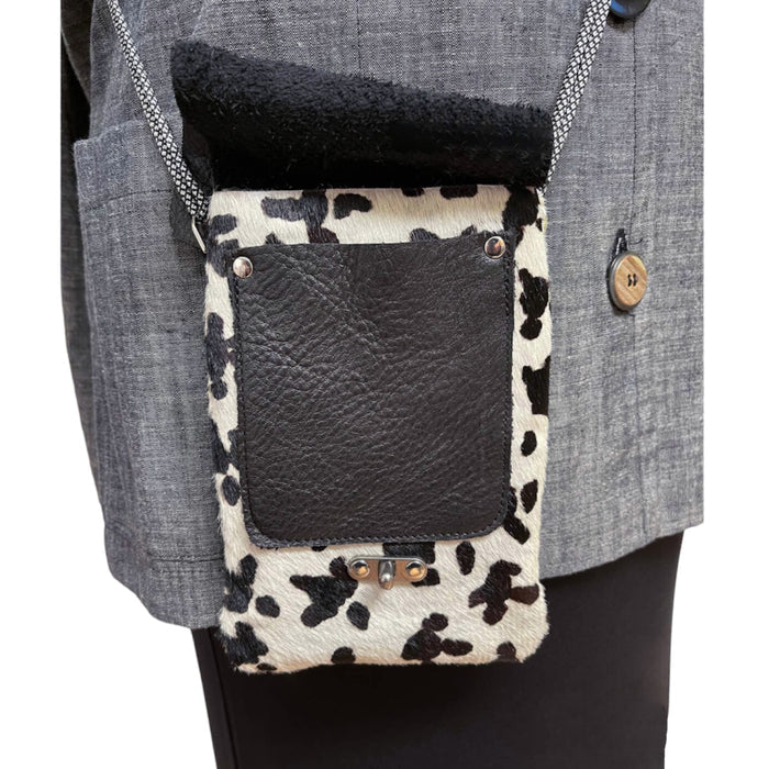 Small crossbody bag with cow print pattern and black leather flap worn over a gray coat.
