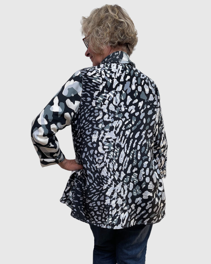 An older woman viewed from the back wearing a black and white abstract patterned button-up shirt and dark blue jeans, set against a solid light gray background.