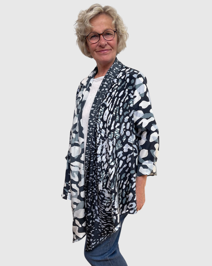 A woman with short gray hair and glasses wearing a black, white, and gray abstract patterned cardigan over a white top and blue jeans, standing against a light gray background.
