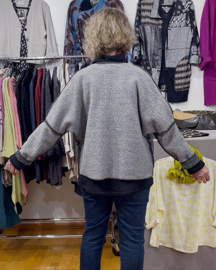 Person wearing a gray sweater and blue jeans in a clothing store with racks of clothes and a table with yellow fabric.